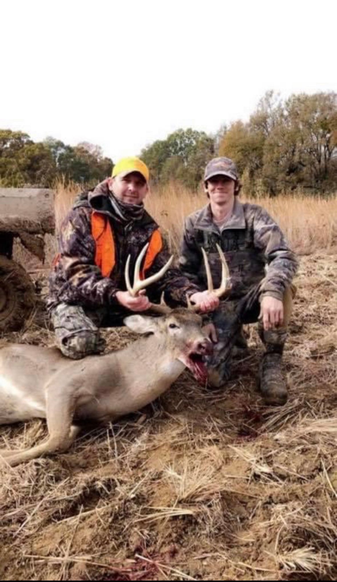 Two hunters kneeling beside a deer in a field, one holding antlers, with a dead goose nearby.