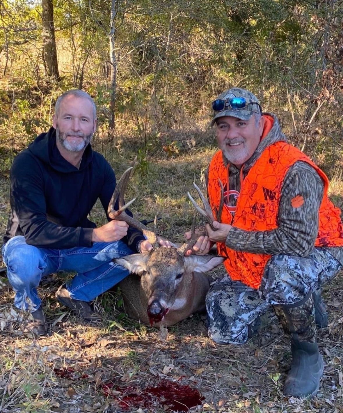 Two hunters kneel in the woods beside a deer, one in orange camouflage, holding the antlers.