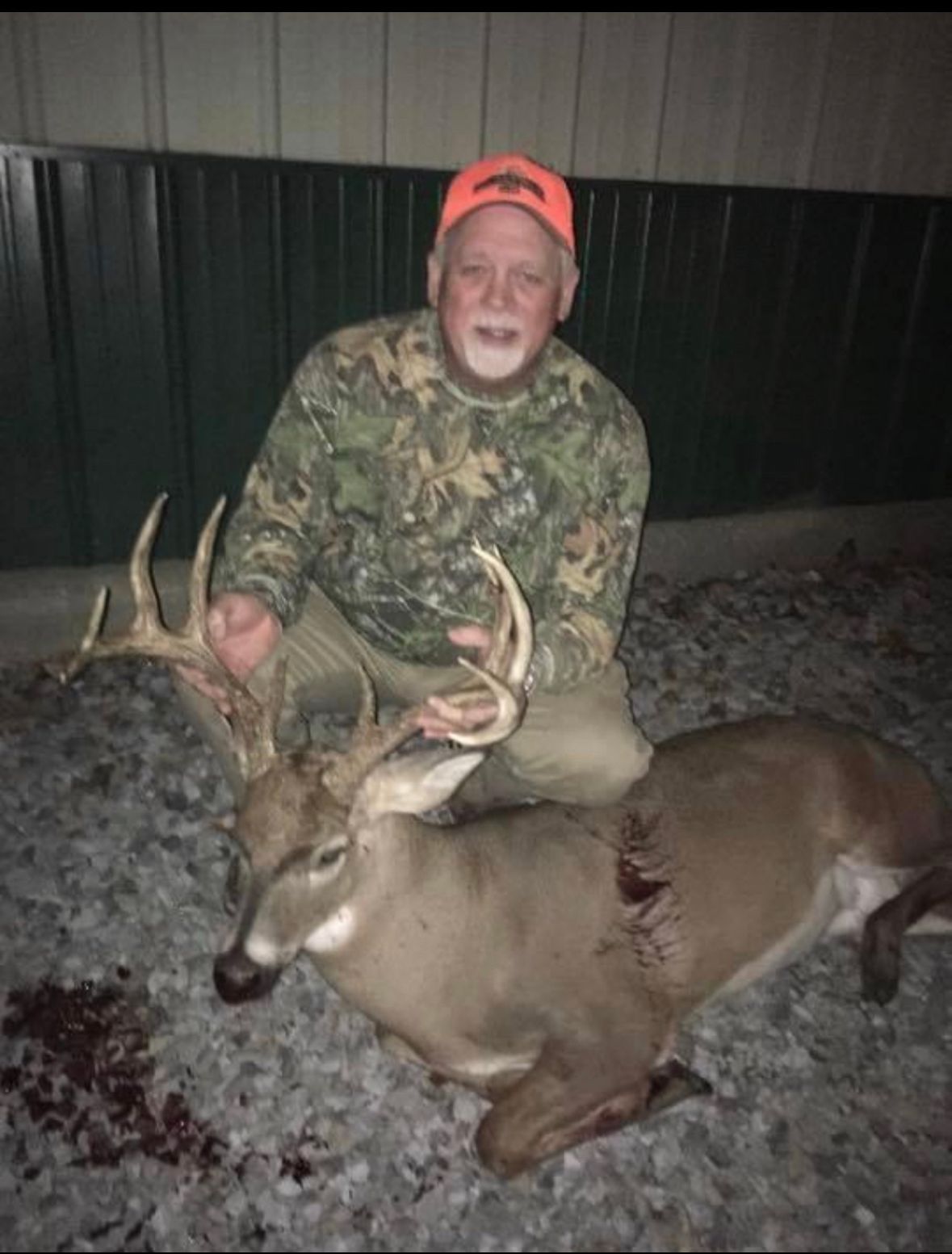 Hunter kneeling beside a large deer with antlers at night, wearing camouflage and an orange cap.
