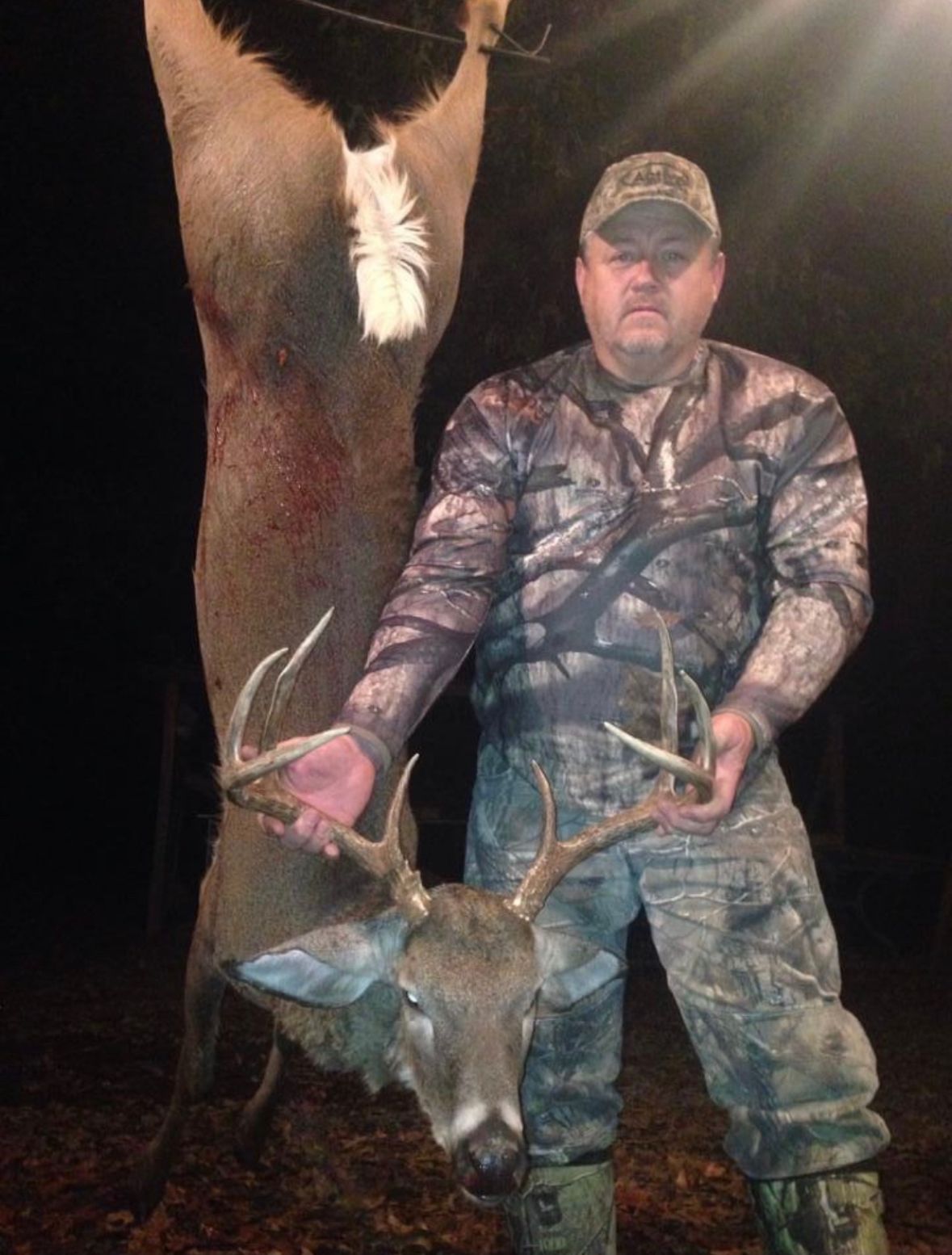 Hunter posing with a deer at night, wearing camouflage and holding the antlers.