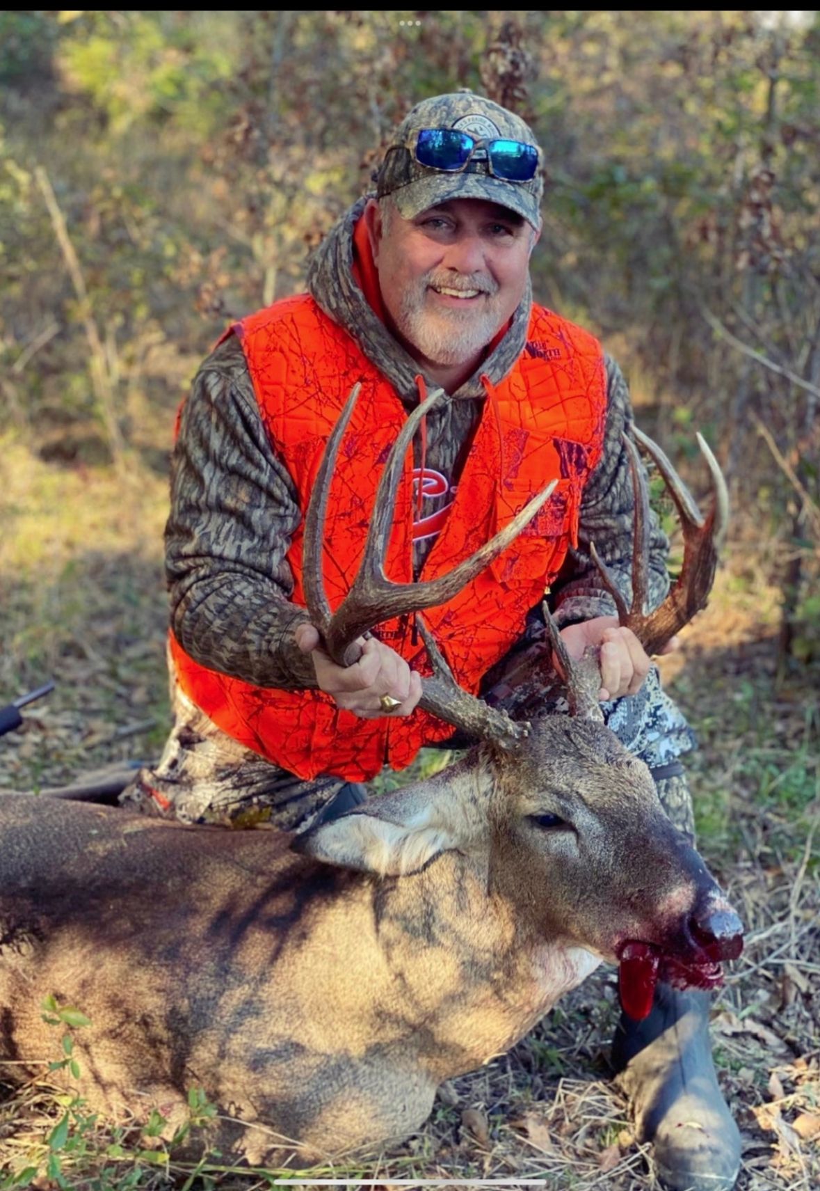 Man in bright orange vest posing with a deer in a wooded area
