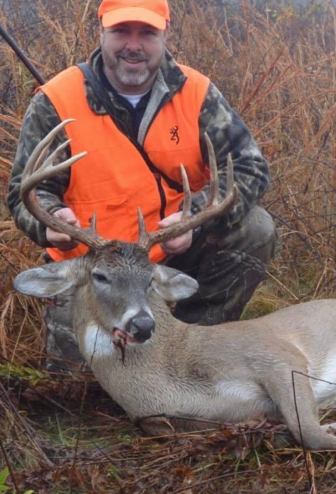 Hunter in orange vest and cap kneels beside a large buck in a brushy field.