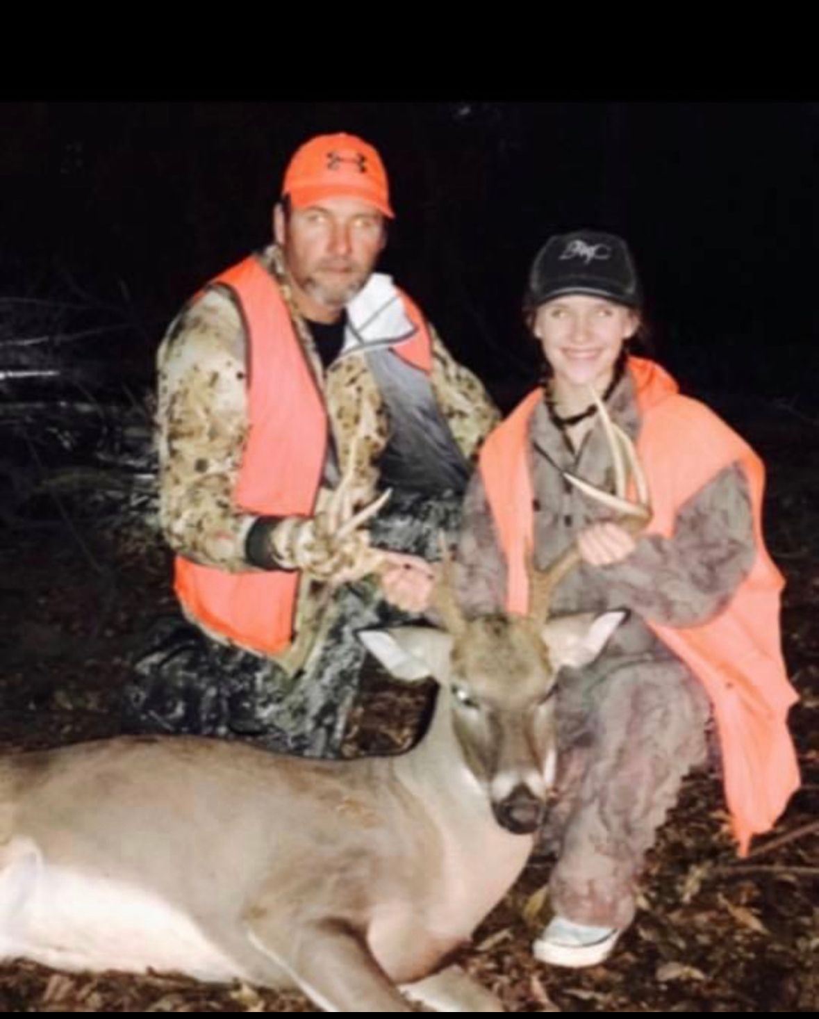 Two hunters in orange vests posing with a deer at night in the woods