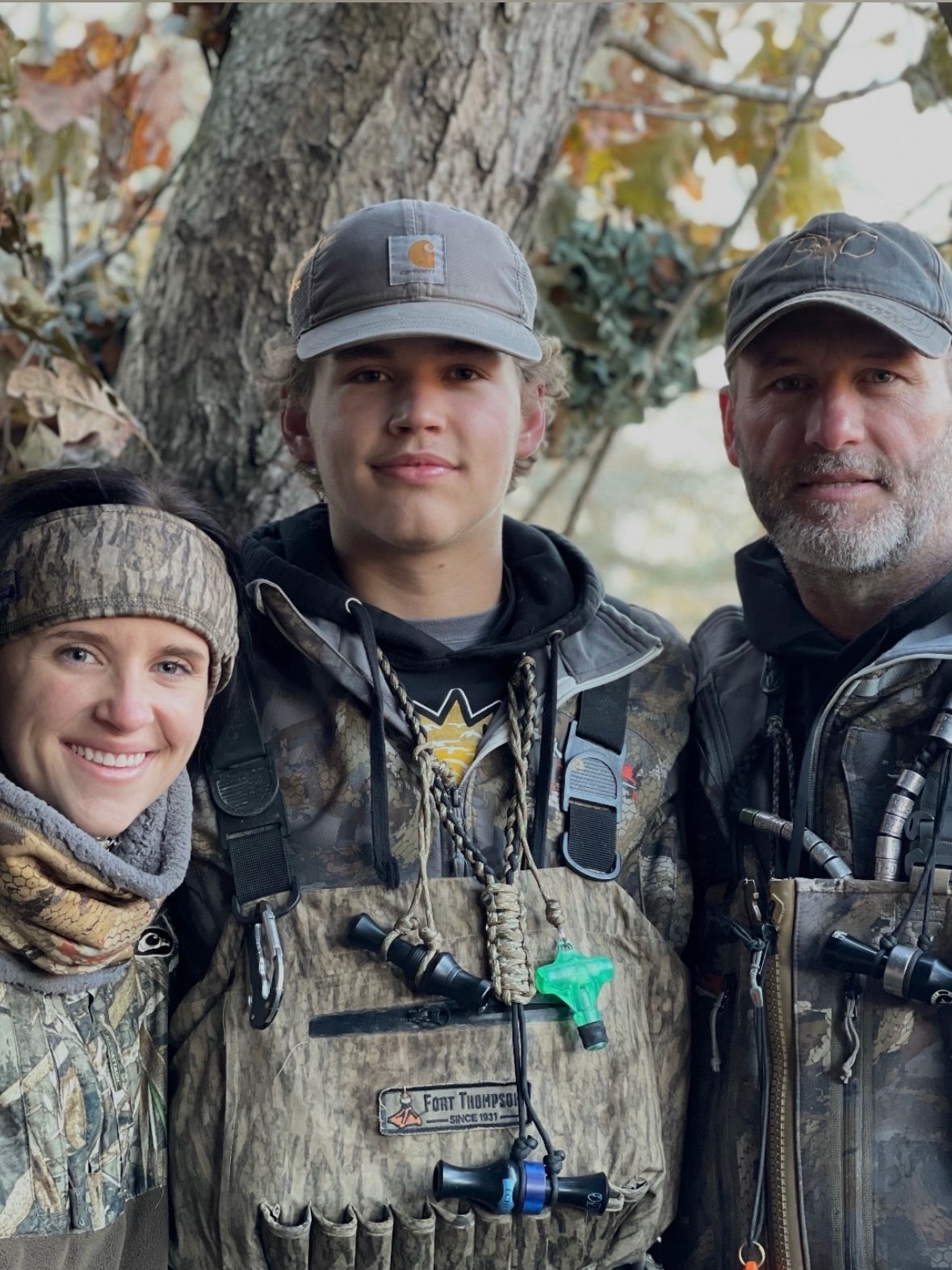 Three hunters in camo standing outdoors among autumn trees, two smiling at the camera.