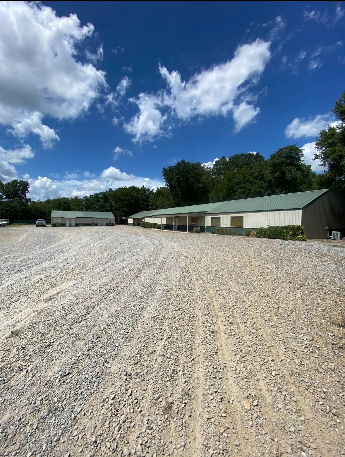 Gravel lot beside long green-roofed buildings under a bright blue sky with clouds