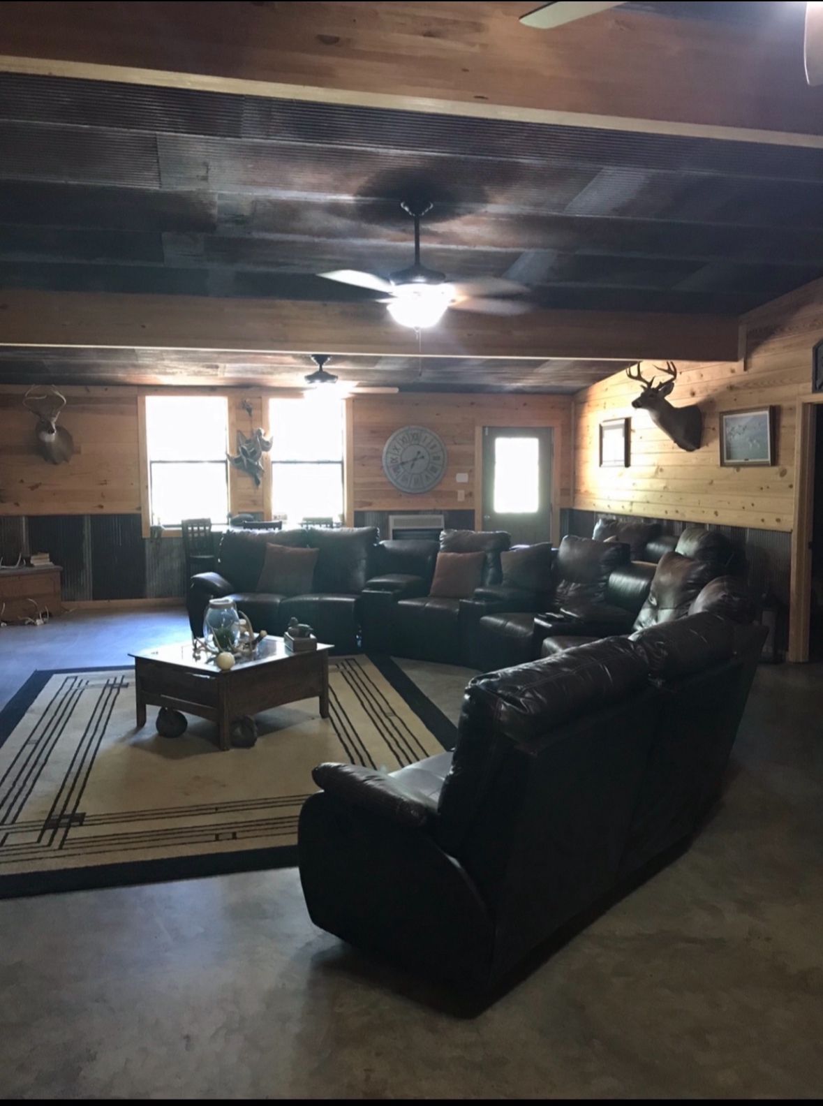 Cozy rustic living room with leather sofas, coffee table, patterned rug, and a ceiling fan under wood-paneled walls