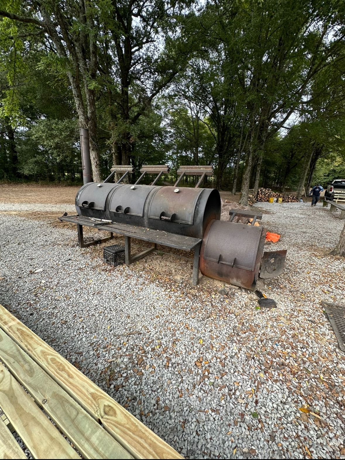 Large black metal smoker grill on gravel in a wooded outdoor area