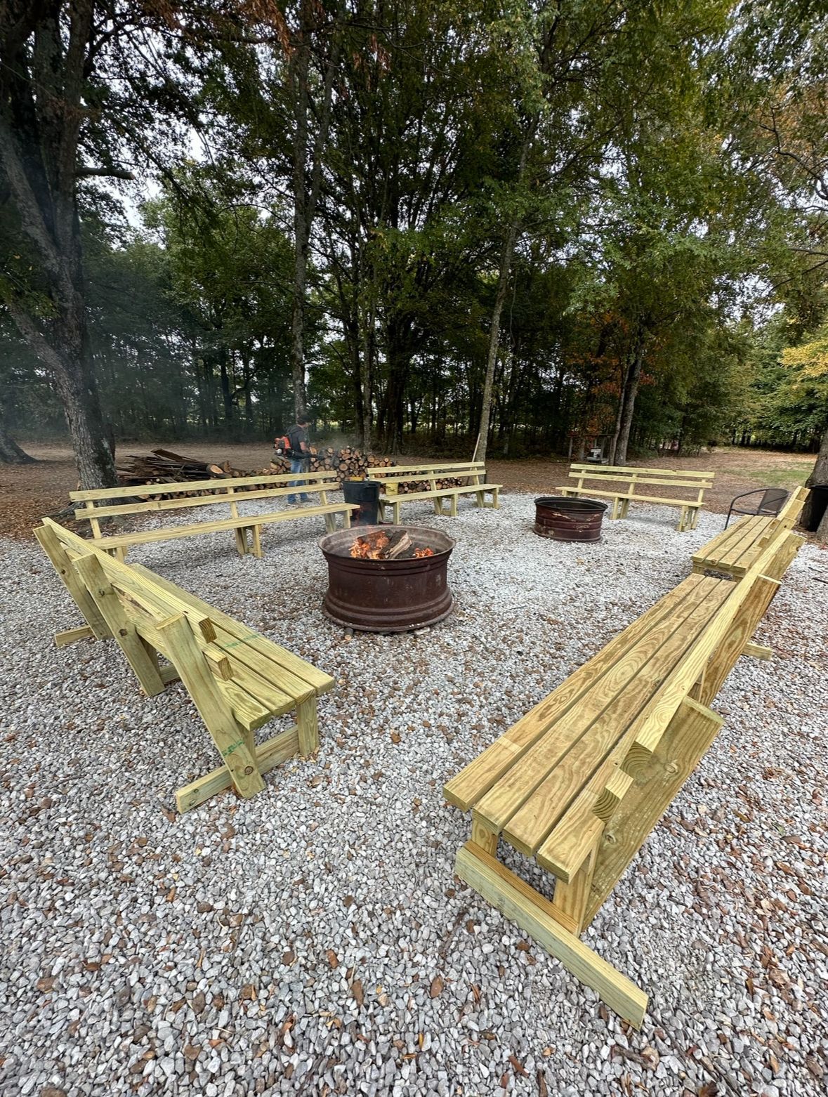 Outdoor gravel fire pit area with wooden benches around two metal fire rings under trees