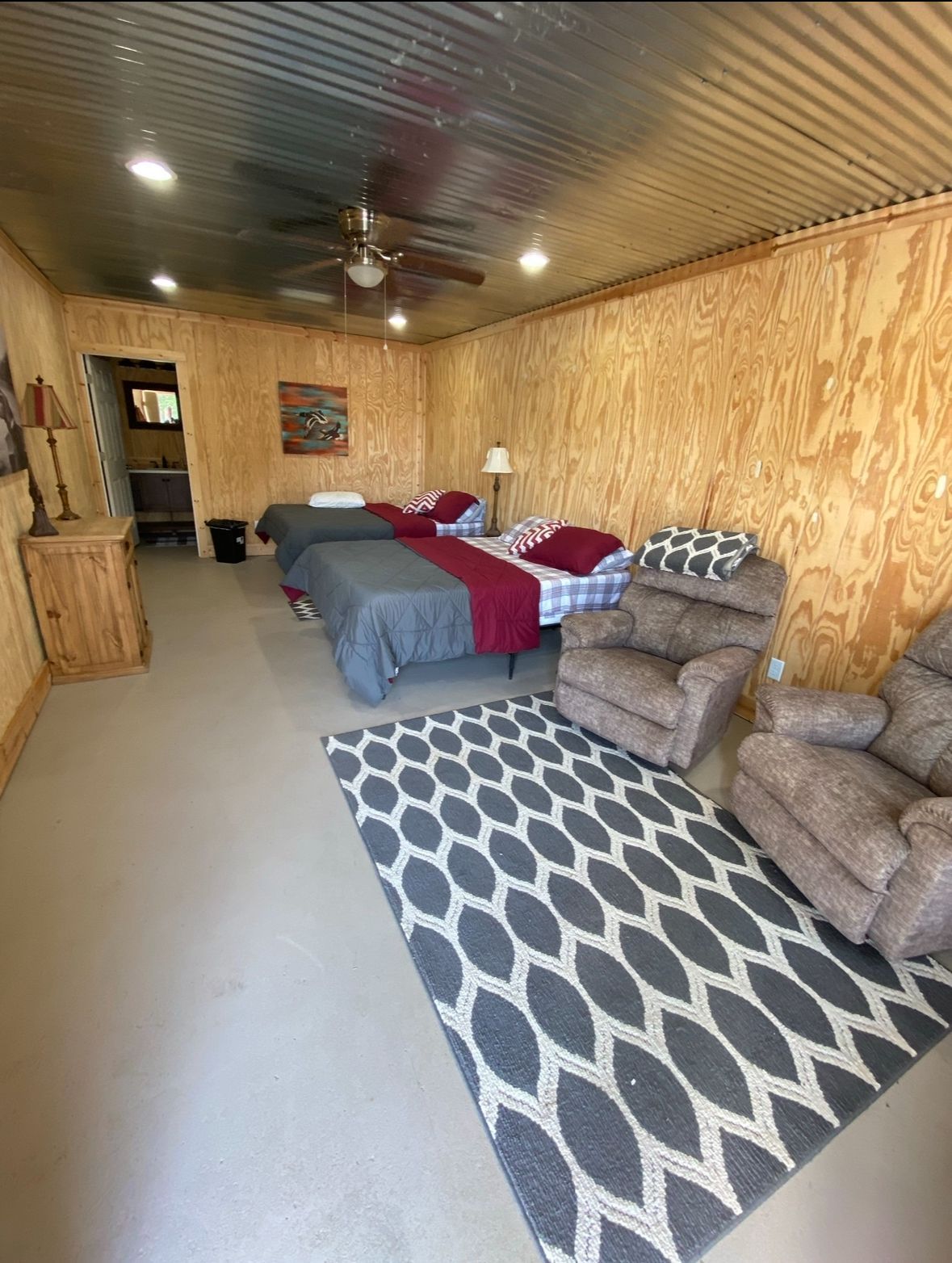Cozy bedroom with wood-paneled walls, two beds, armchairs, and a patterned rug under ceiling lights.