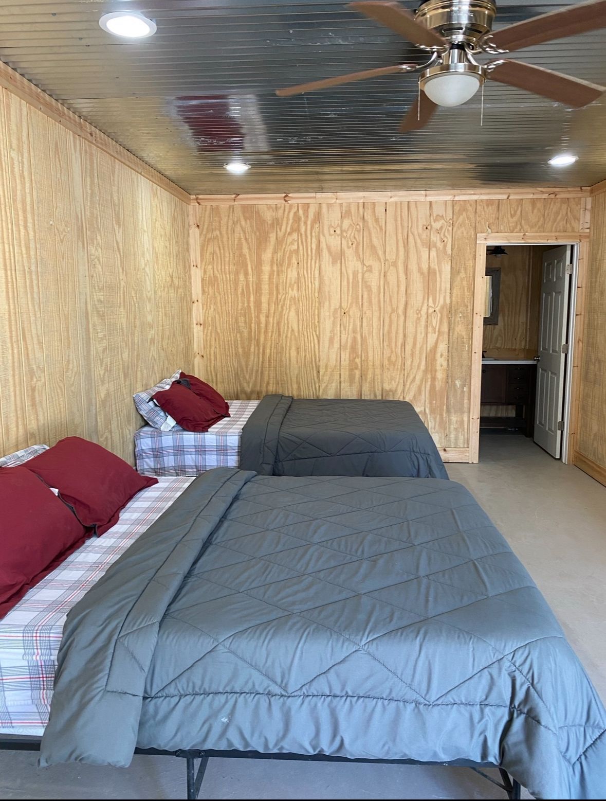 Small wood-paneled bedroom with two beds, gray comforters, red pillows, and a ceiling fan.