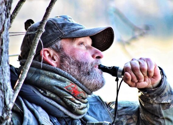 Man in a cap blowing a whistle outdoors, wearing a gray scarf and jacket.