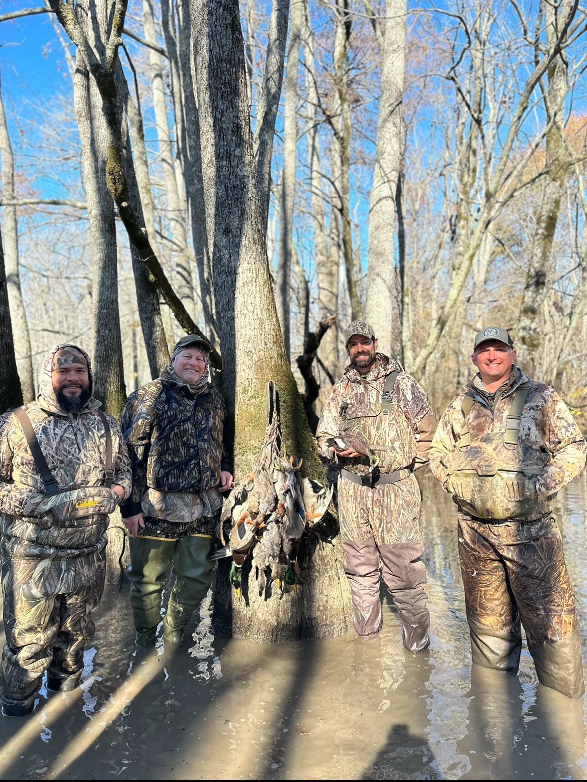 Four people in camouflage waders standing in a flooded wooded area, posing with hunting gear.