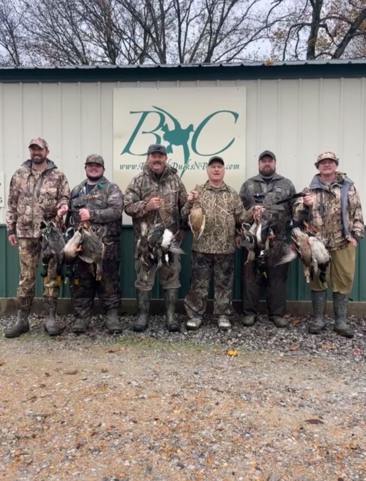 Six hunters in camouflage pose holding harvested birds in front of a BMC sign.