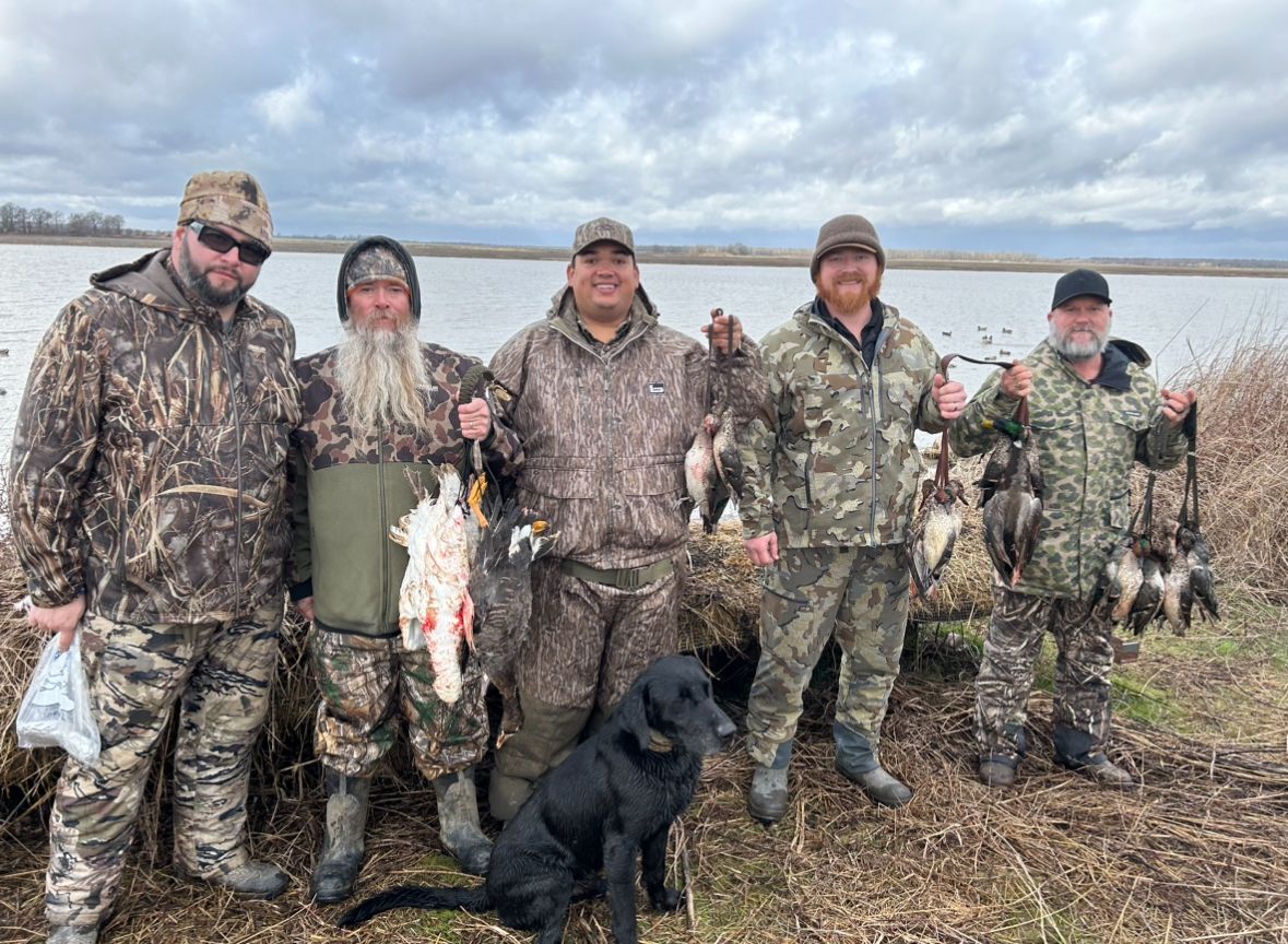 Five people in camouflage gear posing with ducks by a marsh, with a black dog in front