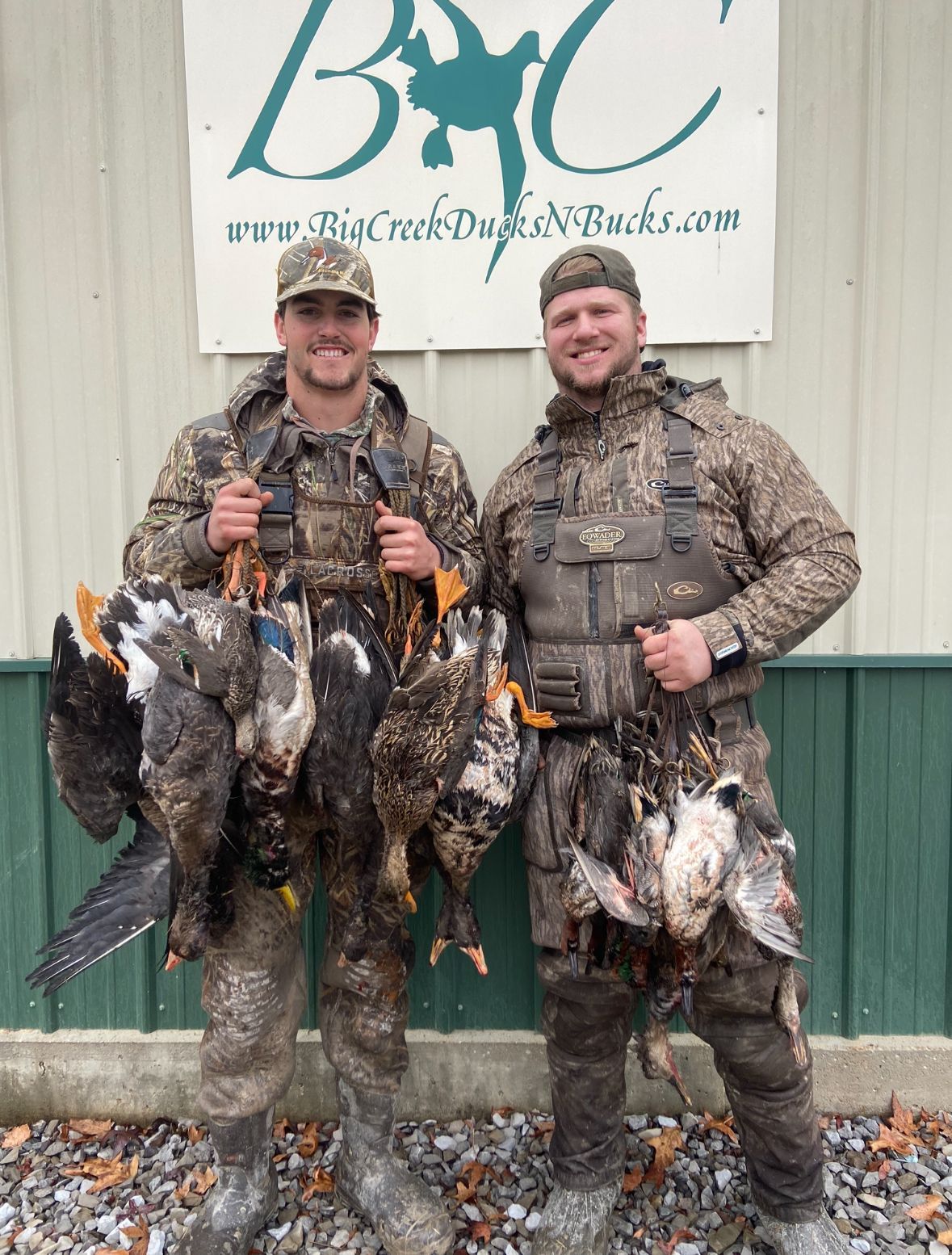 Two hunters in camo holding several ducks in front of a building sign.
