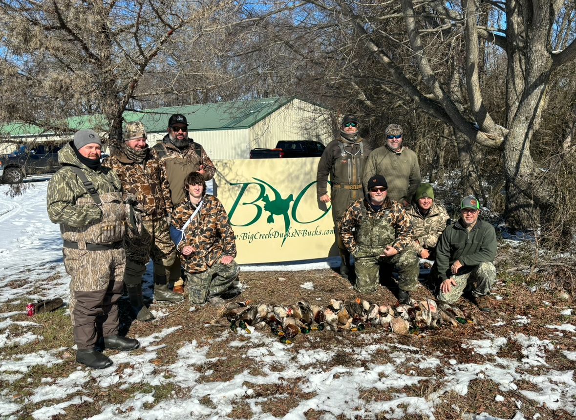 Group in winter camouflage posing beside a tan shelter with green logo in a snowy wooded area