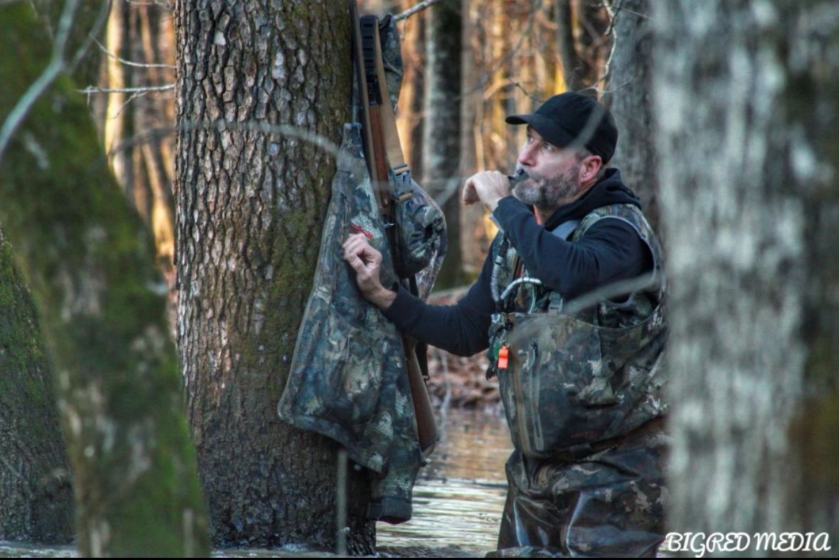 Camouflaged hunter standing in a flooded forest, holding a tree and drinking from a cup