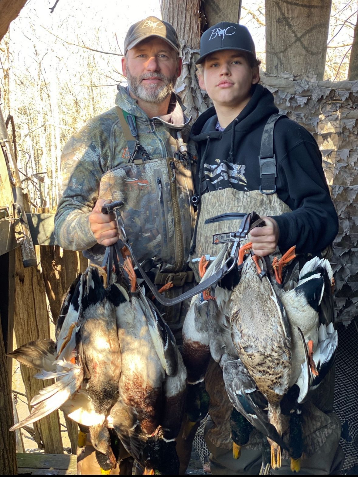 Two hunters standing in woods, holding several harvested ducks by the necks