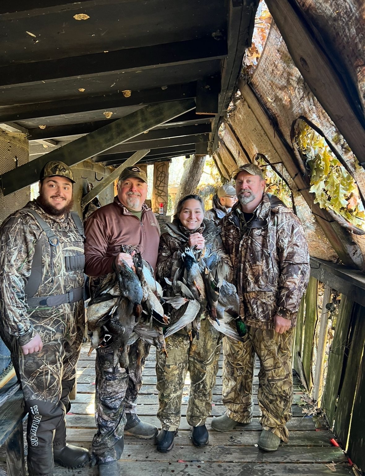 Four hunters in camo pose inside a duck blind holding hunted birds.