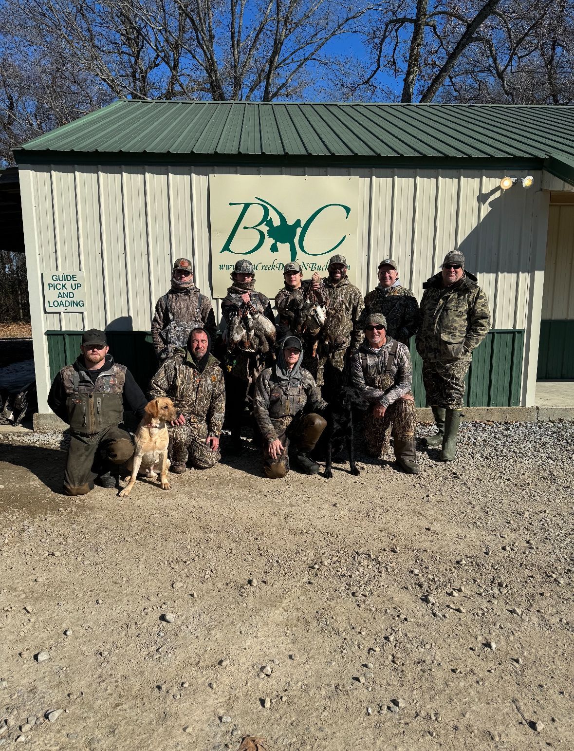 Group in camouflage posing with a dog outside a beige building with a green roof.