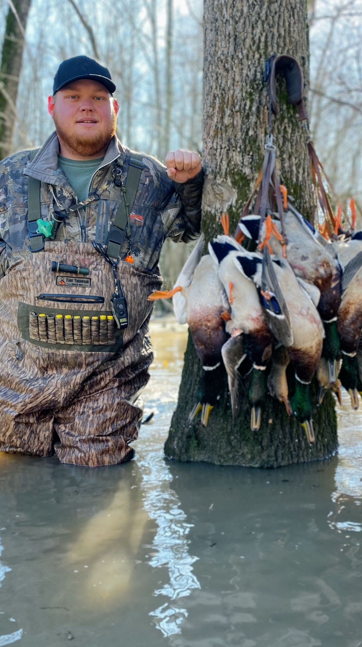 Hunter in camouflage beside a tree with several ducks hanging on display in shallow water.