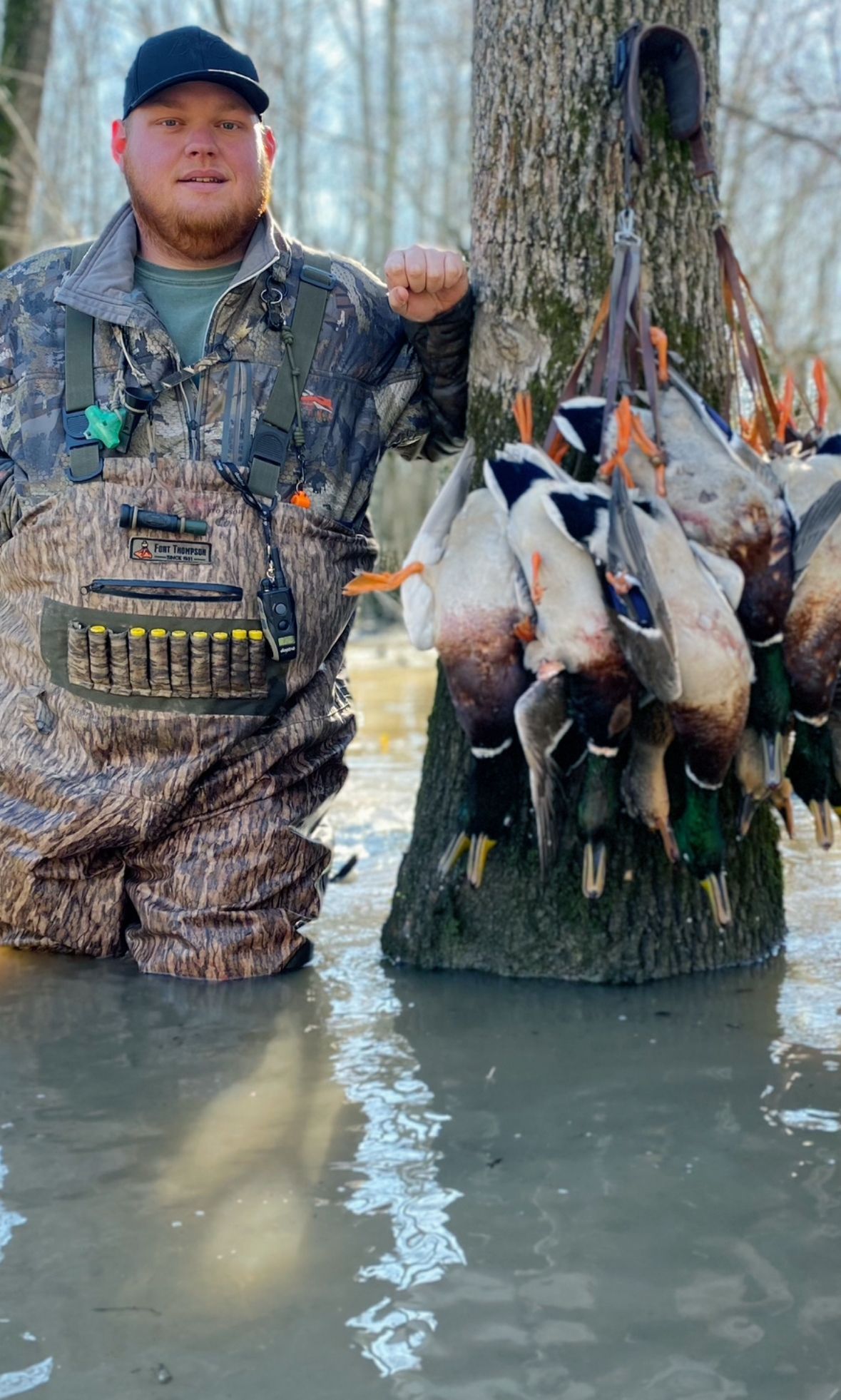 Hunter in camouflage stands in shallow water beside a tree, holding a large stringer of ducks.