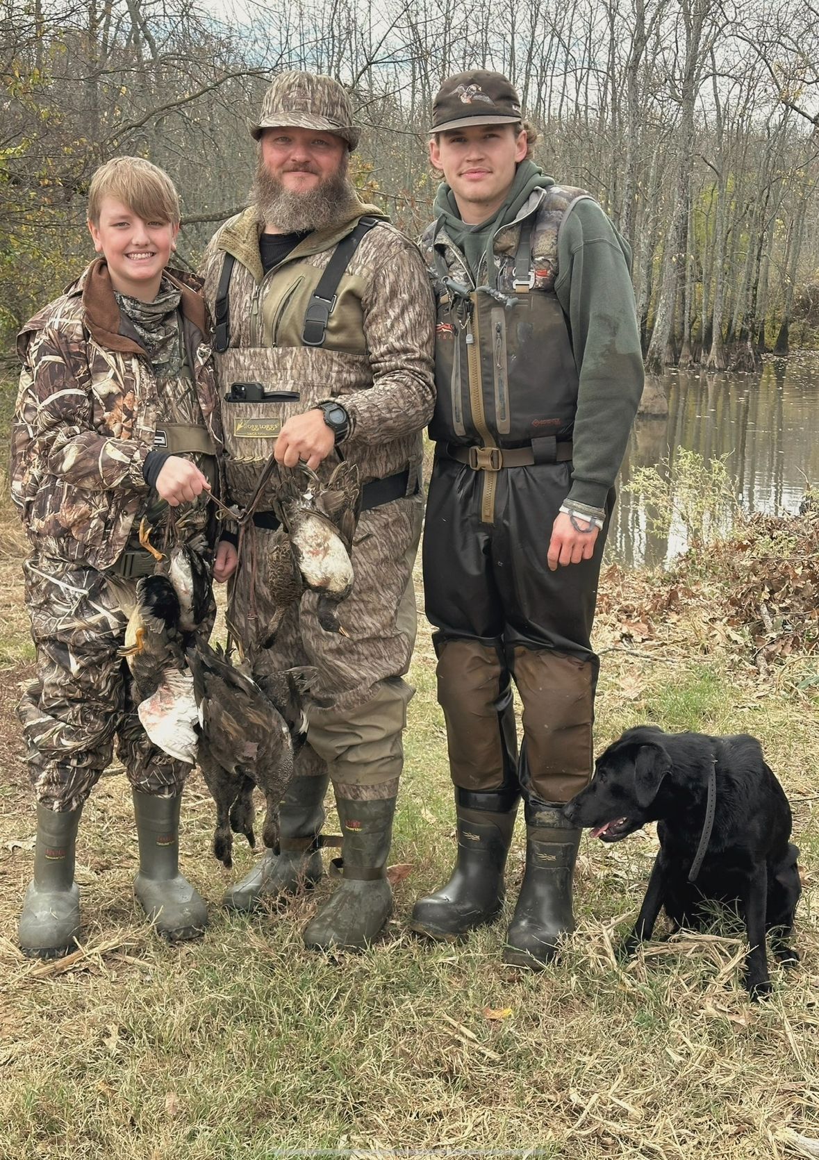 Three people in camo waders pose with a black dog by a flooded marsh.
