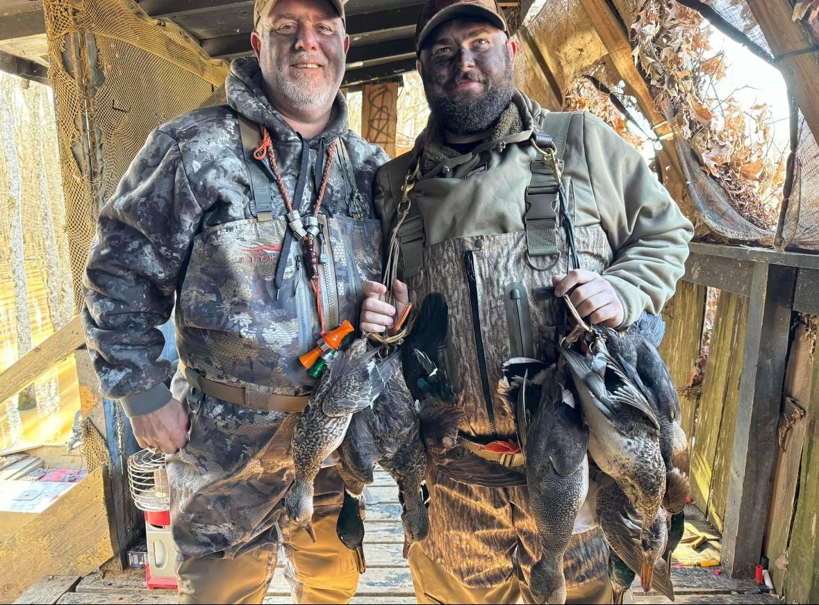 Two hunters in camo stand in a blind, smiling and holding two dead ducks.