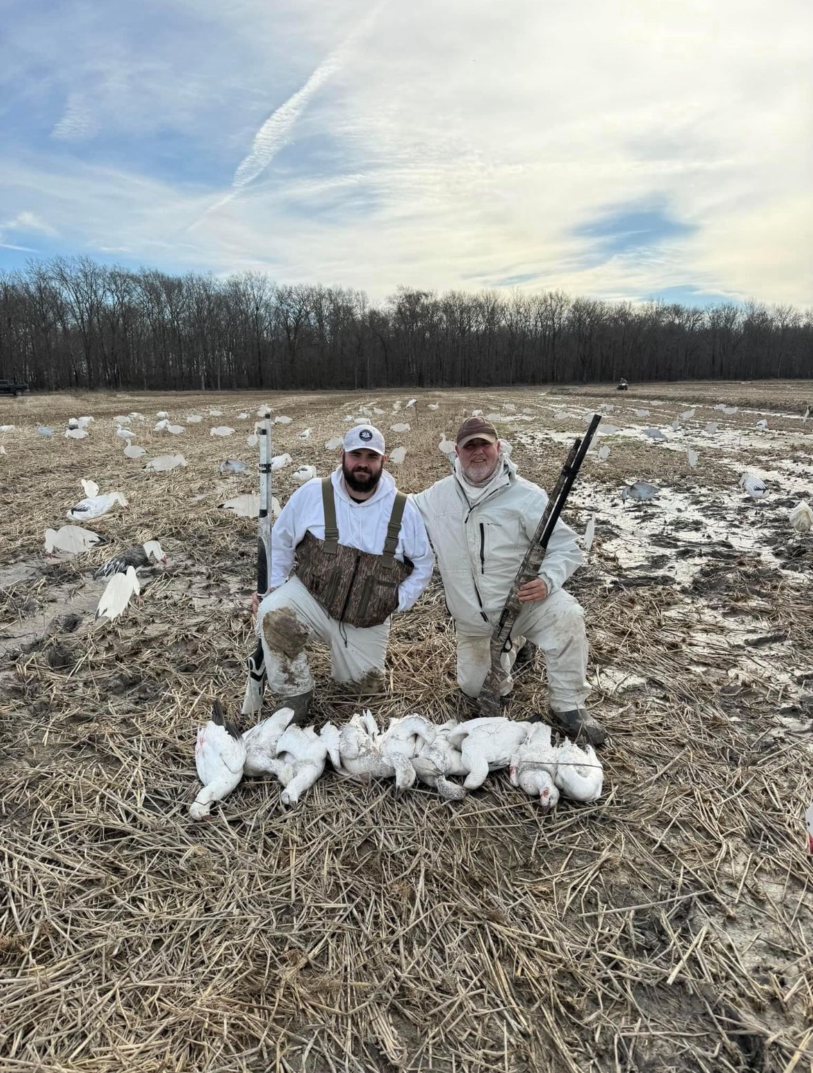 Two hunters kneel in a snowy field with a row of harvested ducks and a shotgun at their side.