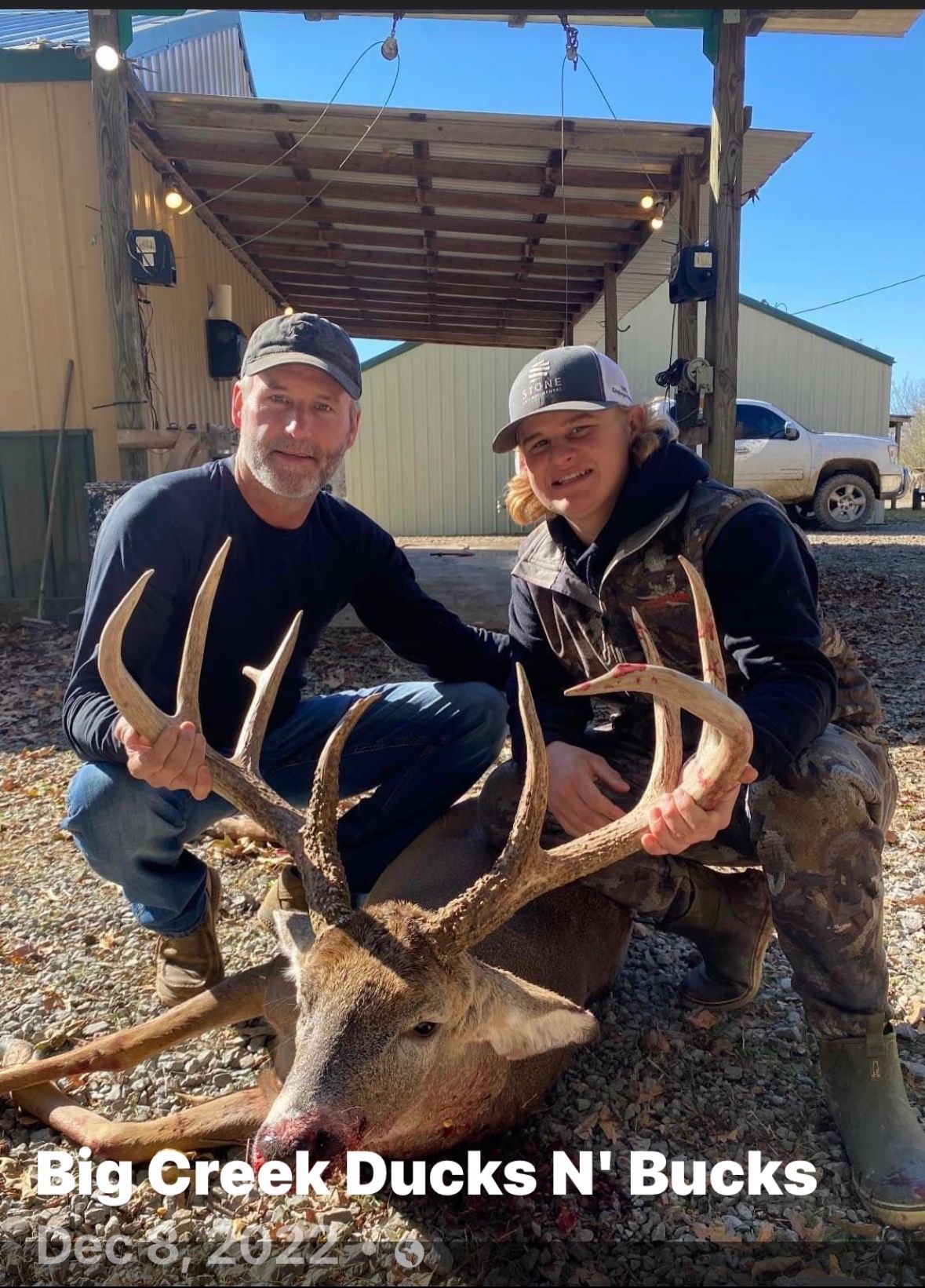 Two people kneel behind a large deer buck with big antlers outside a rustic building.