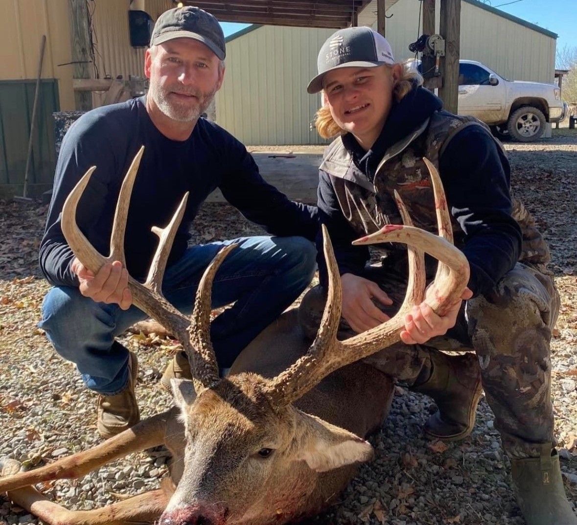 Two hunters kneel beside a large deer with antlers outside a metal building.