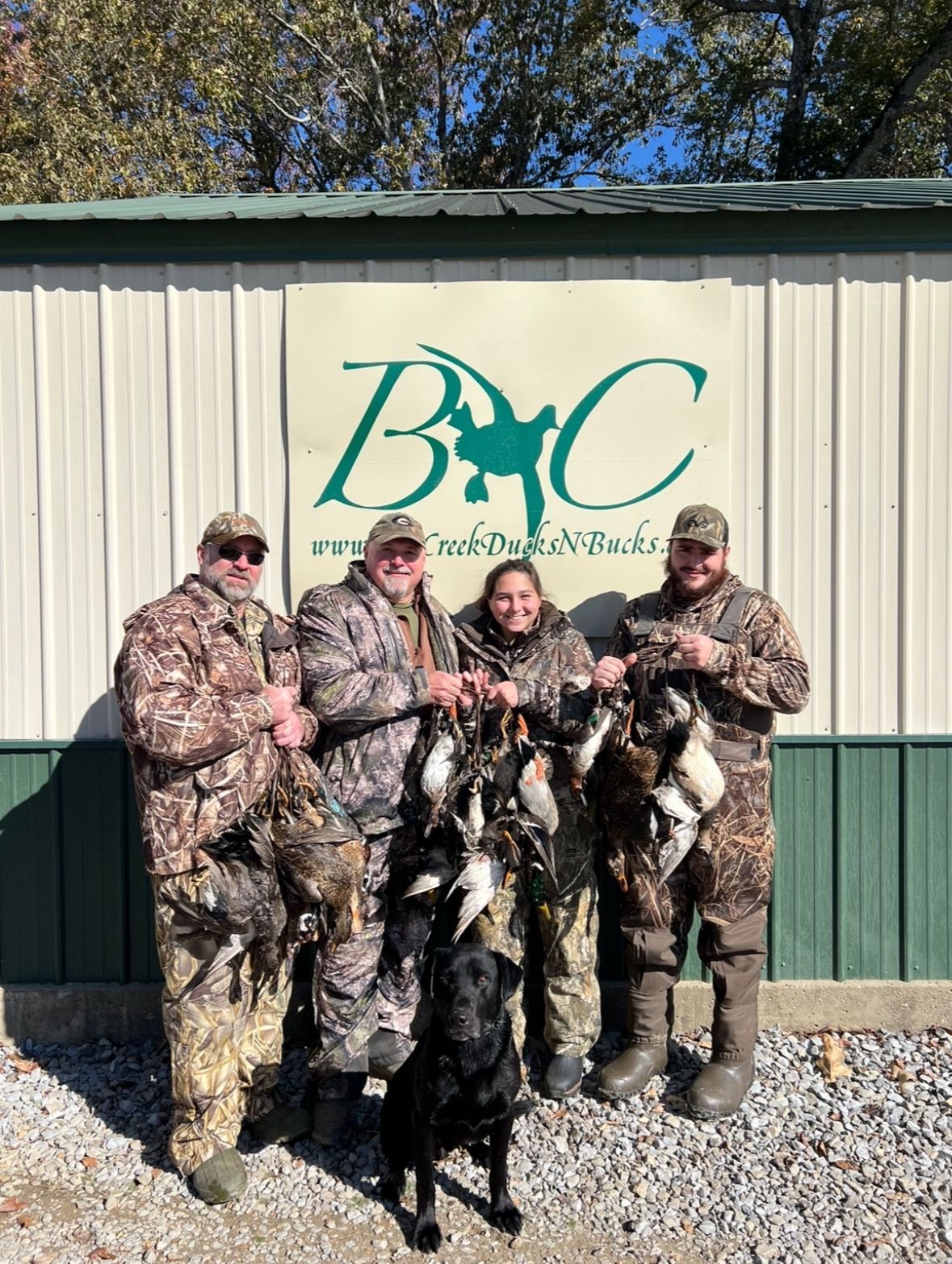 Five people holding hunted birds in camouflage in front of a B&C building sign