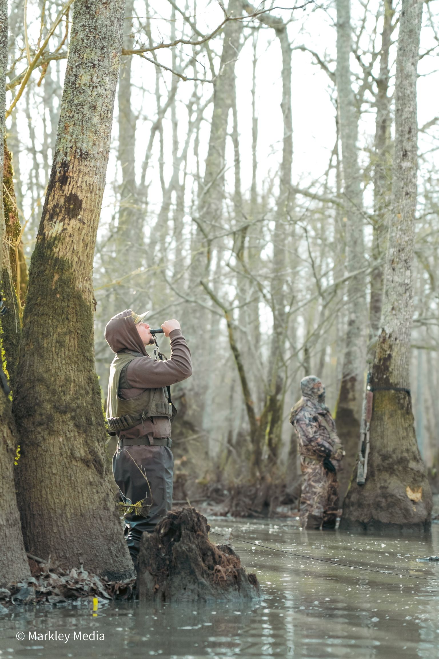Two people in a flooded forest, one photographing wildlife among bare trees.