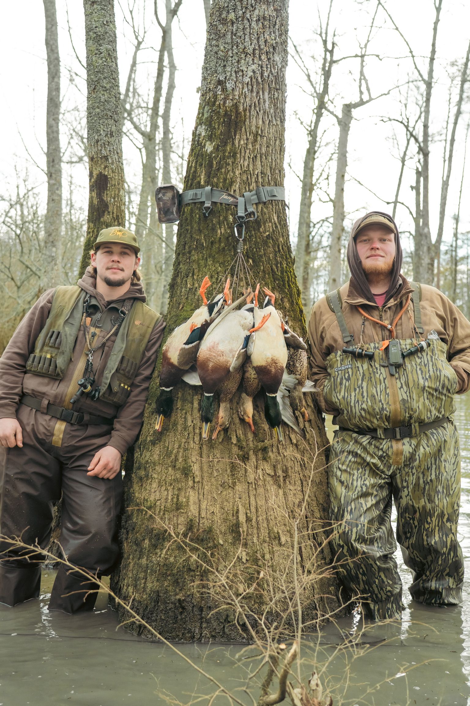 Two people in camo standing in shallow water beside a tree with several ducks hanging from it