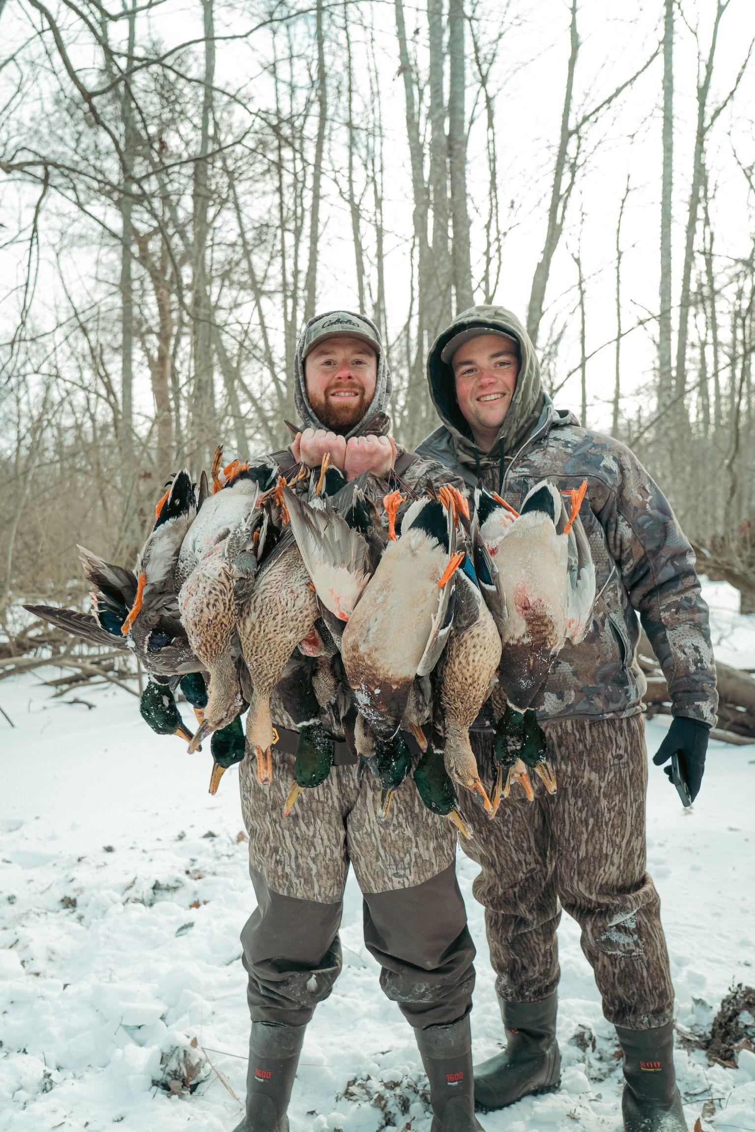 Two hunters in winter gear hold a string of freshly caught birds in a snowy forest