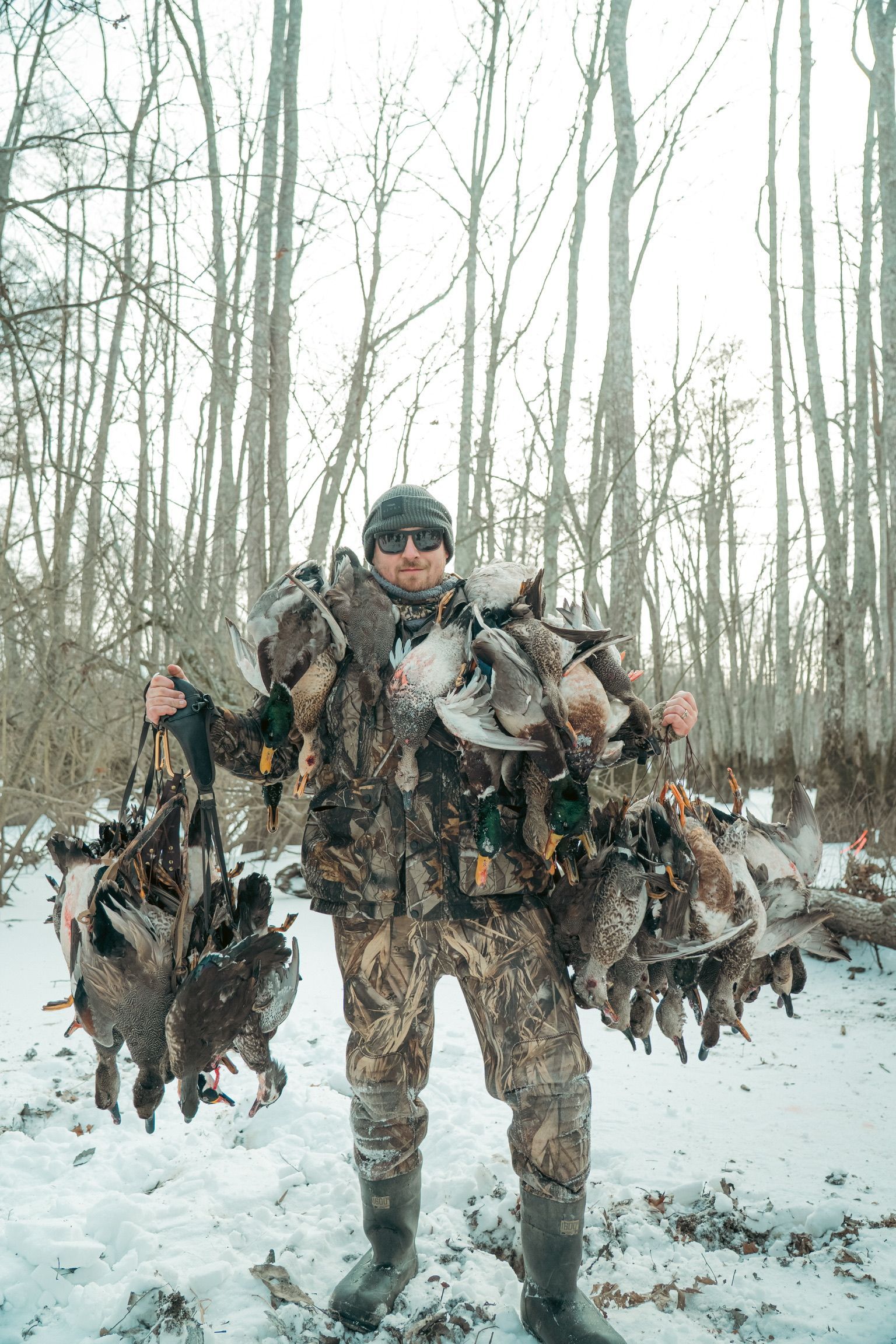 Hunter in camouflage standing in a snowy wooded area, holding several ducks by the feet.