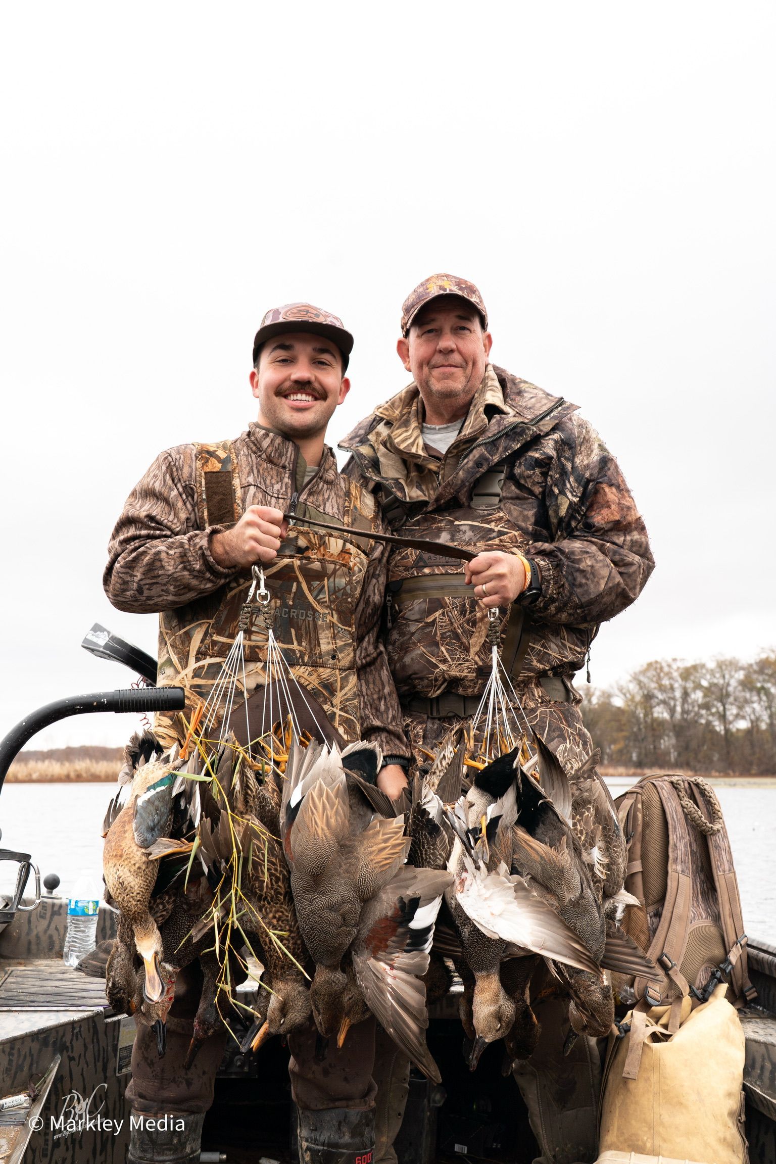 Two hunters in camouflage pose with several ducks beside a boat on a lake.