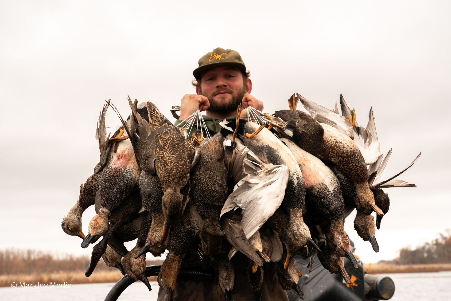 Hunter holding a large bundle of ducks and geese by a lakeshore under an overcast sky
