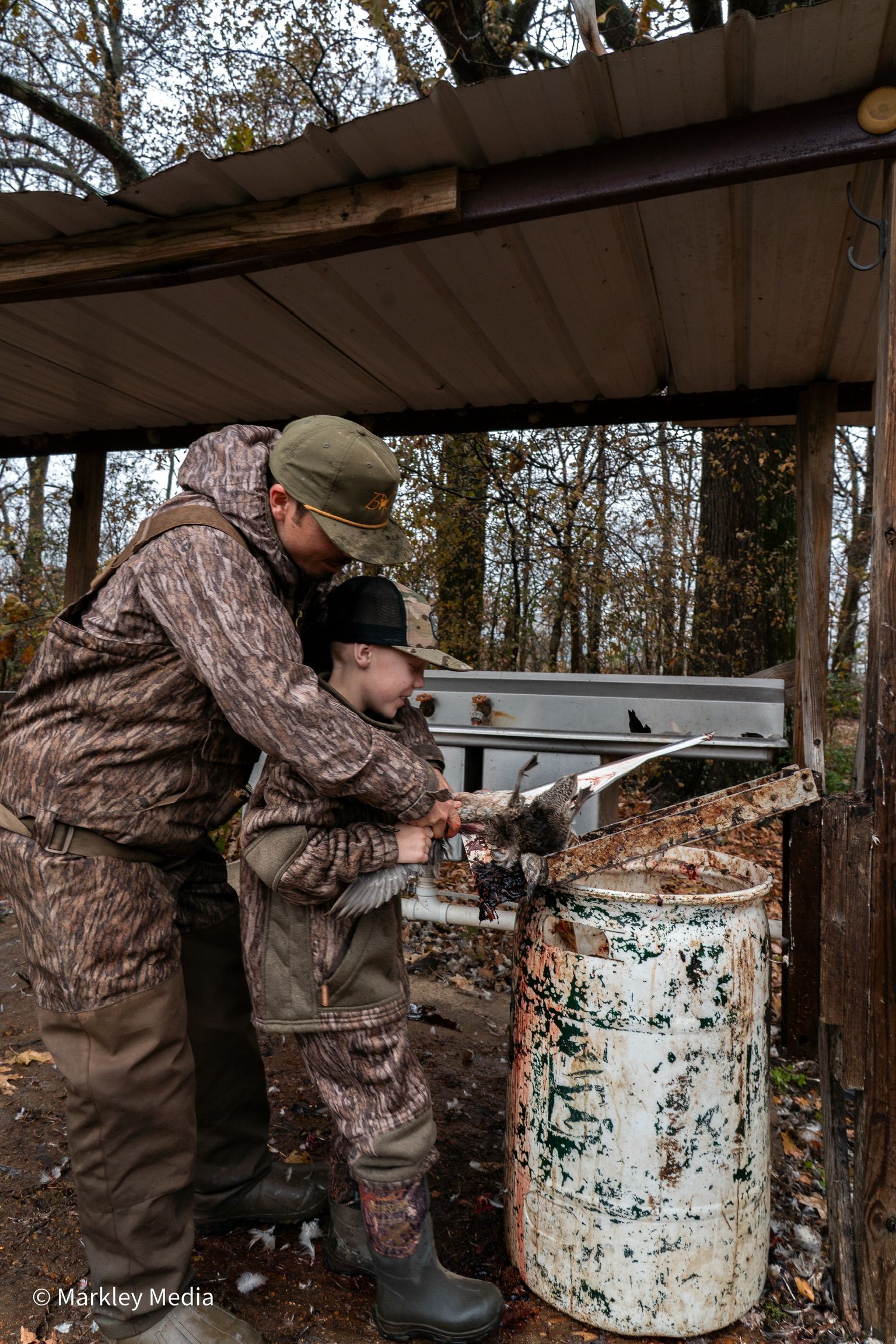 Two people in camouflage coats work at an outdoor metal tank under a shelter in a wooded area.