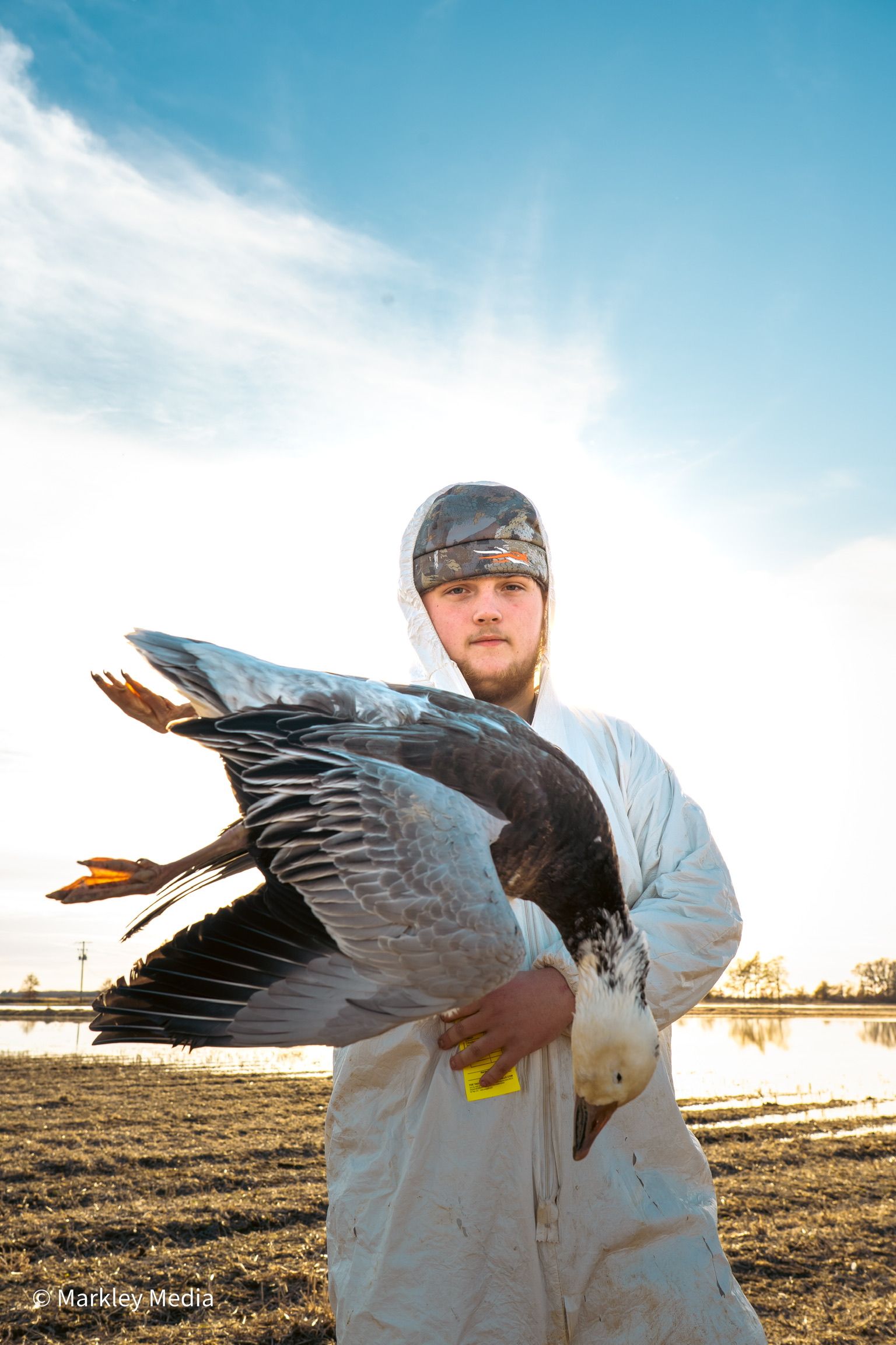 Person in white coveralls holding a large duck outdoors at sunset