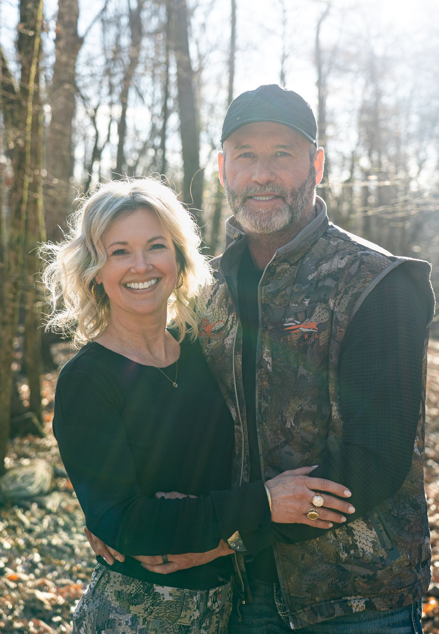 Smiling couple standing outdoors in a wooded area, with one wearing a camouflage vest and cap.