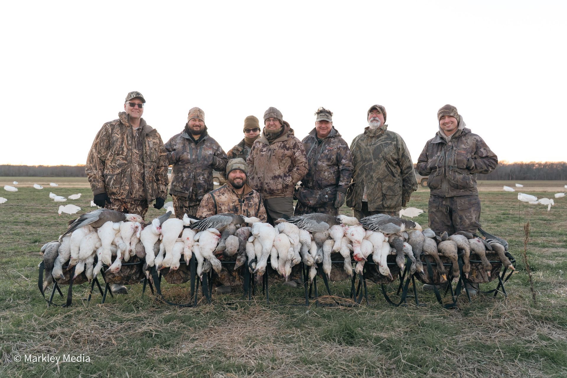 Hunters posing in camouflage with a line of white birds in a field at dusk