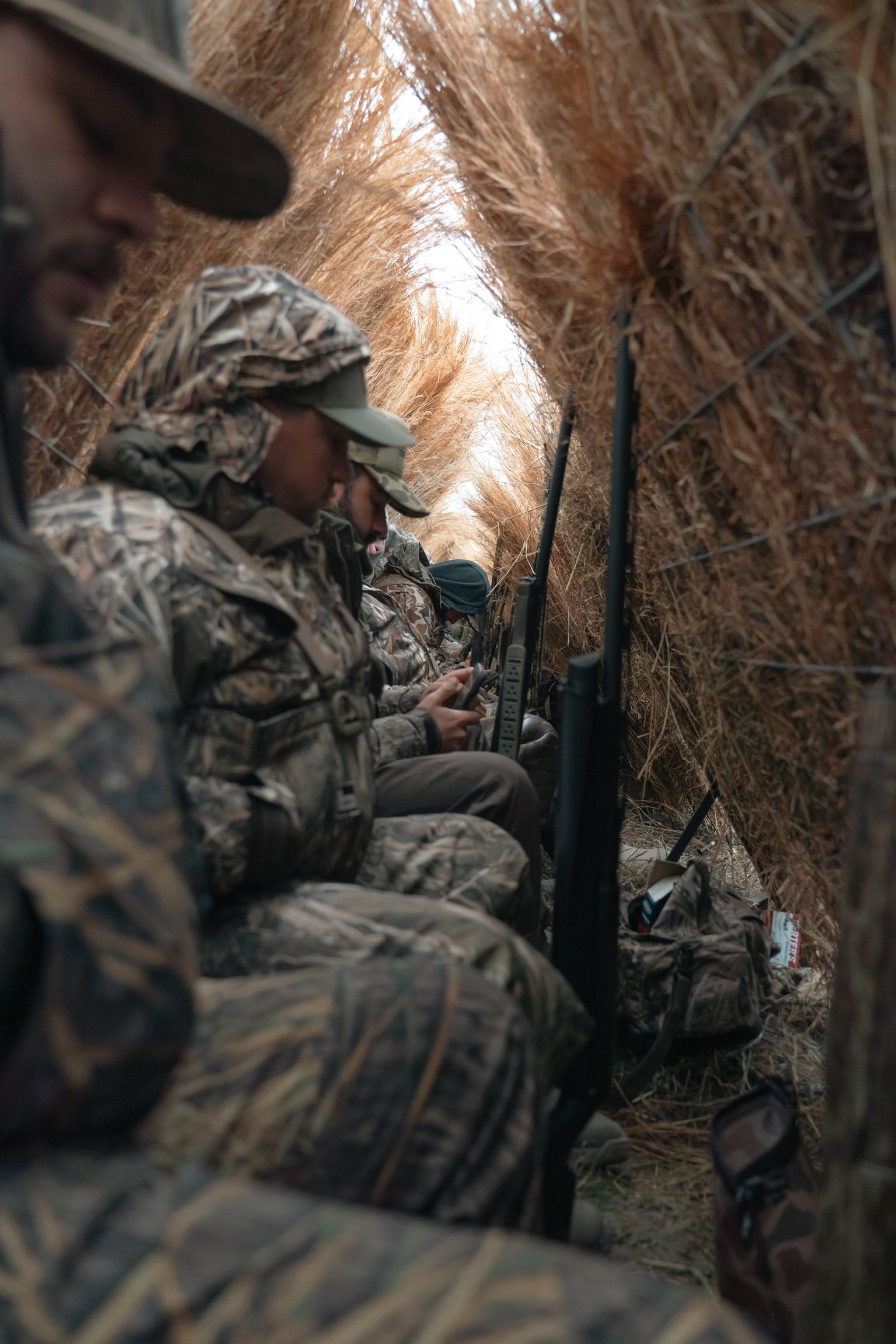 Soldiers in camouflage crouch in a narrow trench lined with dry grass.