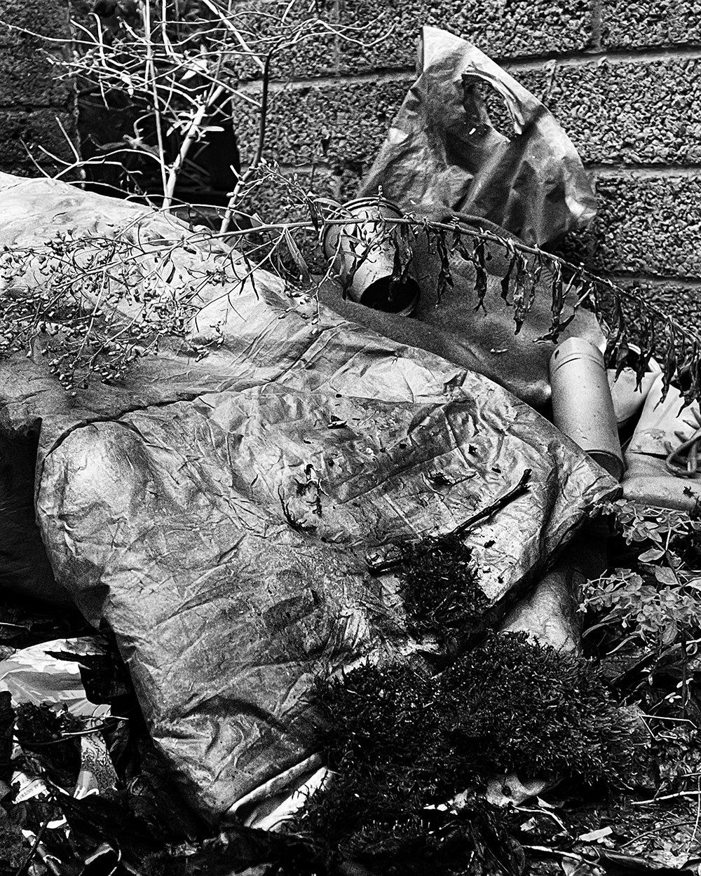 Black-and-white photograph of an empty sleeping bag lying beside weeds, cups, and discarded objects in a neglected laneway