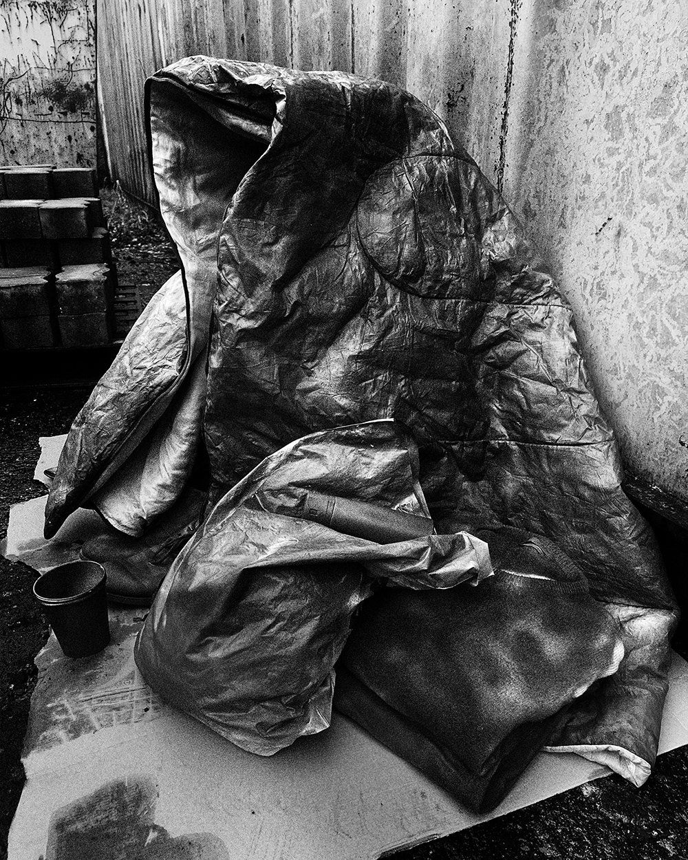 Black-and-white photograph of an empty sleeping bag shaped like a seated body, placed against a wall on a sheet of cardboard
