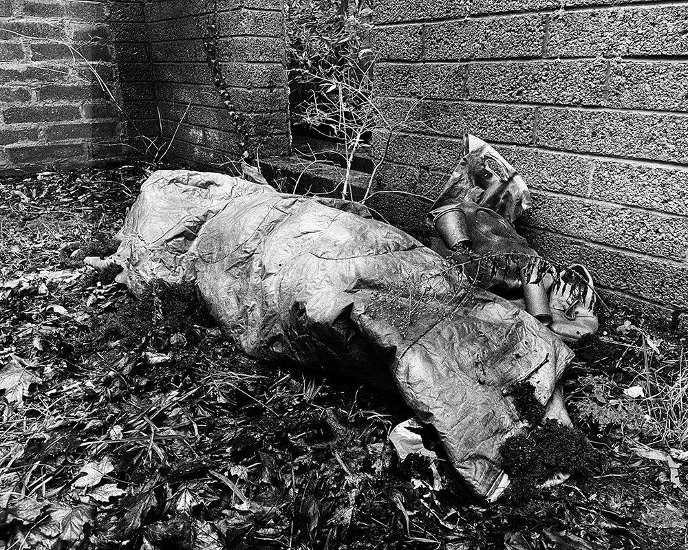 Black-and-white photograph of an empty sleeping bag arranged to resemble a resting body in a neglected alleyway