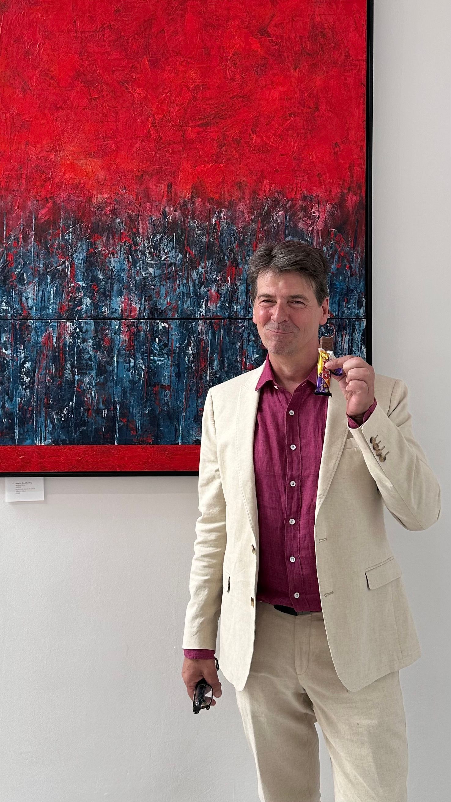 Artist laughing mid-bite of a Cadbury Crunchie  in front of Under a Blood Red Sky at his solo exhibition opening