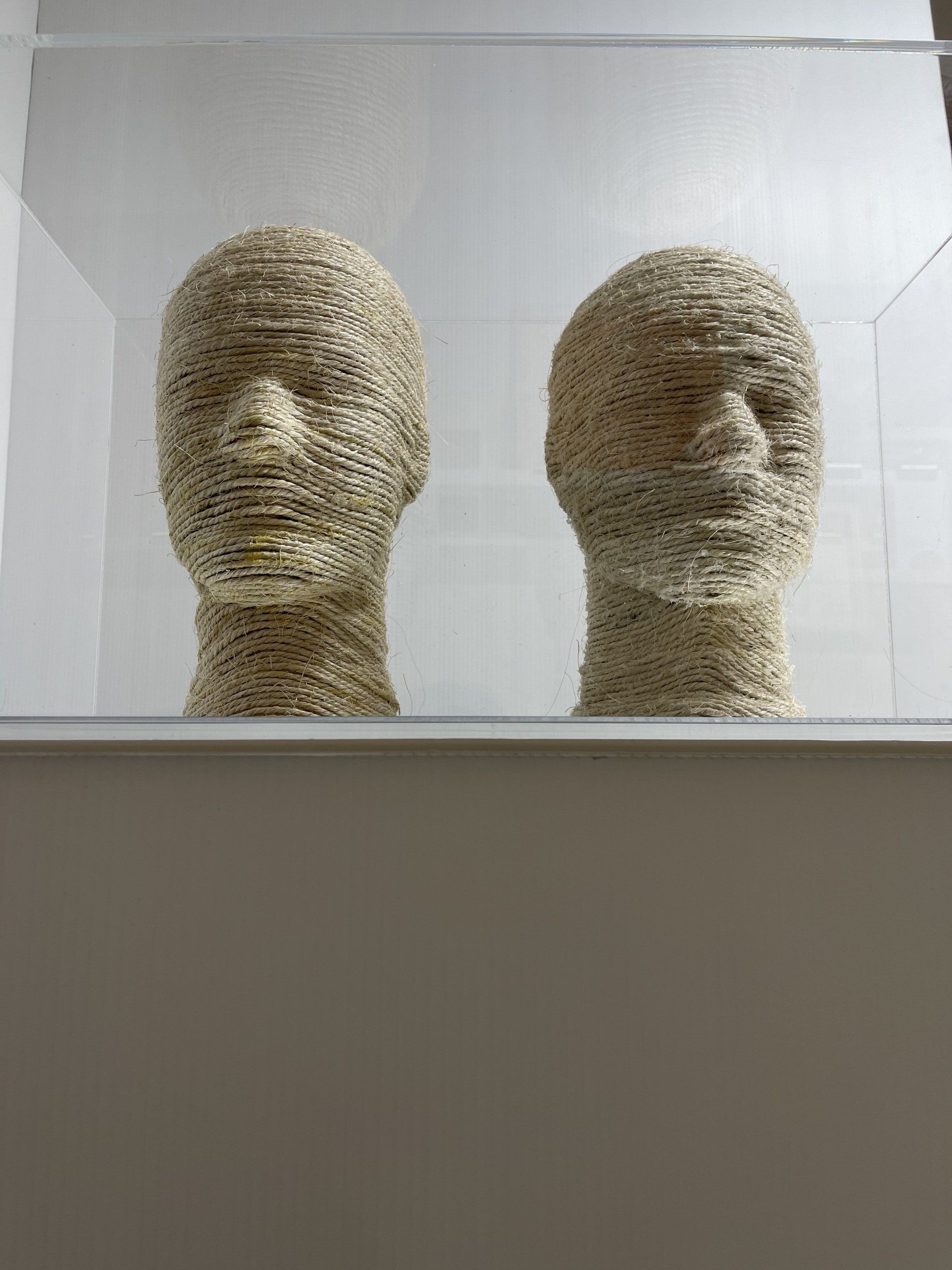 Expressly Vacant, two sisal-wrapped mannequin heads facing forward inside a clear acrylic box with a mirrored base