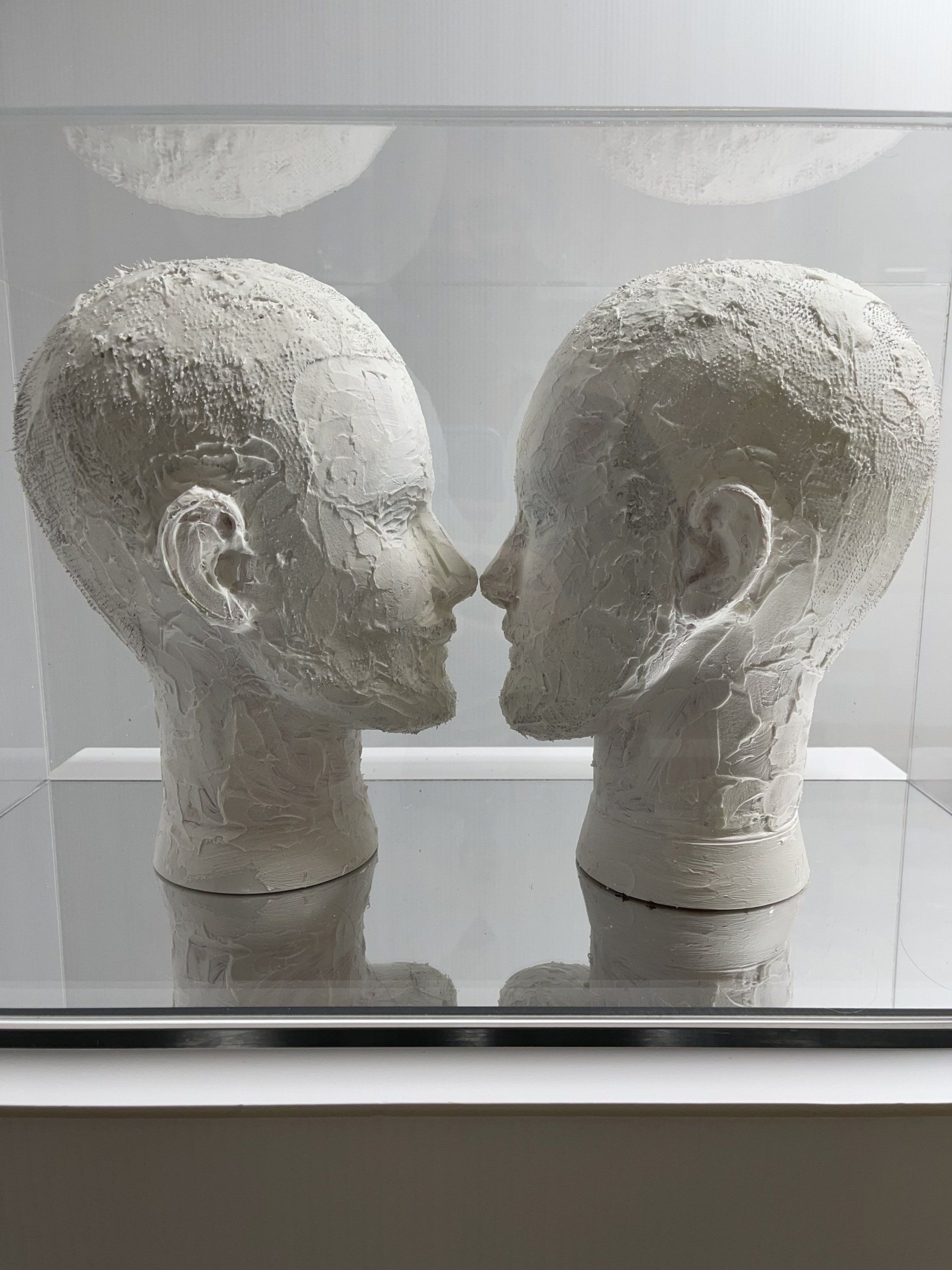 What Next, two plaster-covered mannequin heads facing each other inside a clear acrylic box with a mirrored base
