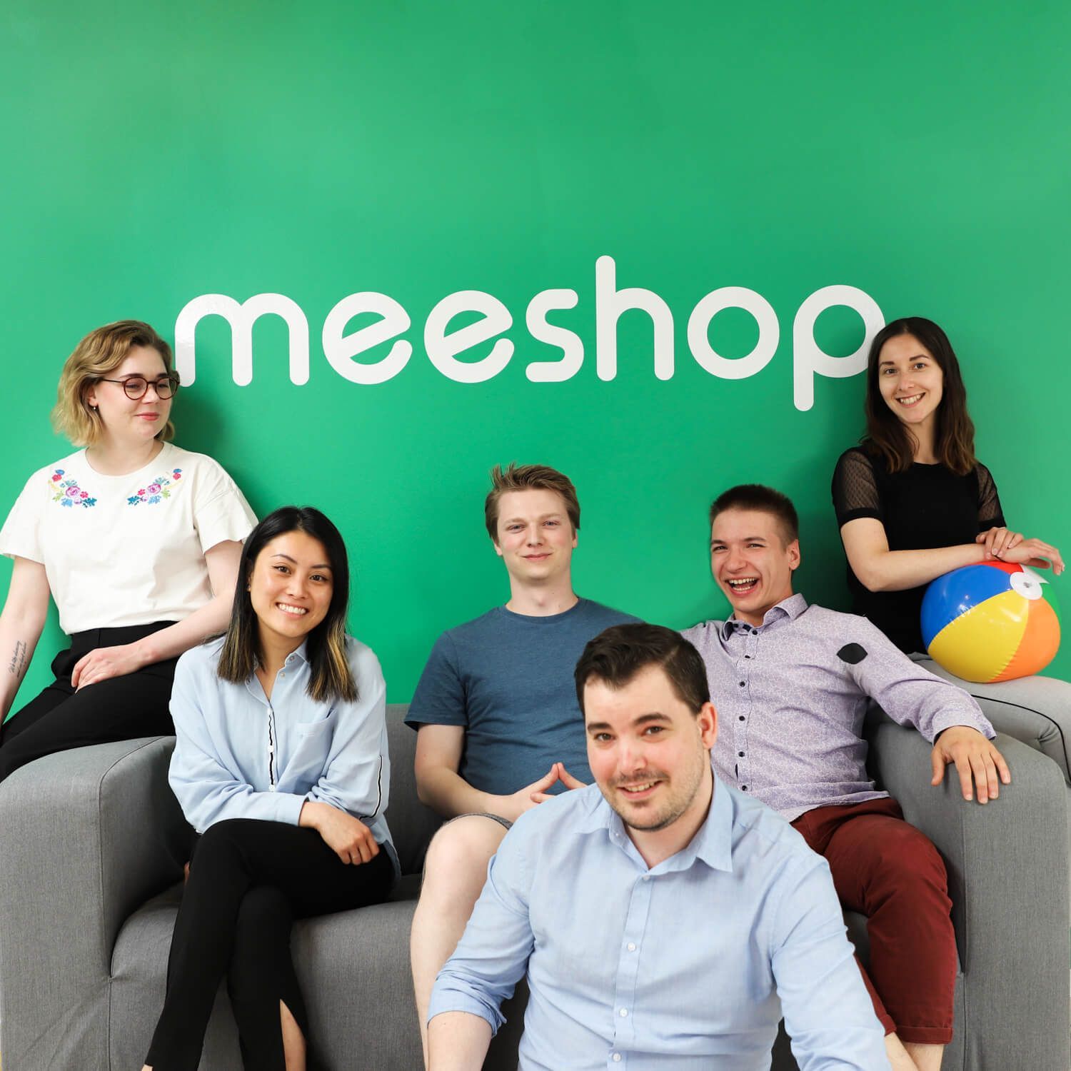 En gruppe mennesker, der sidder på en sofa foran en grøn væg, hvor der står meeshop
